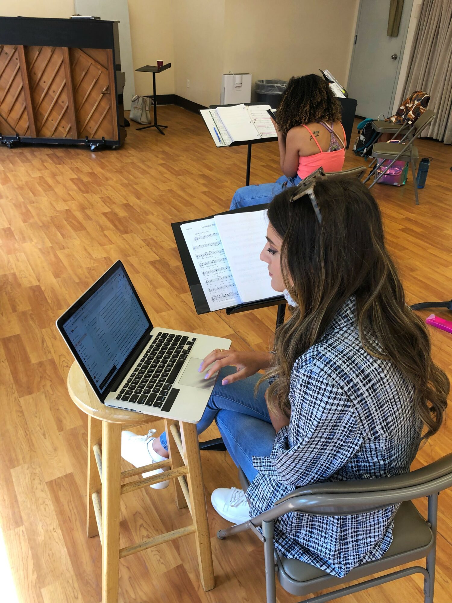 Person working on a laptop at a desk in a room with wooden floor and music stand, with a child reading a book in the background.