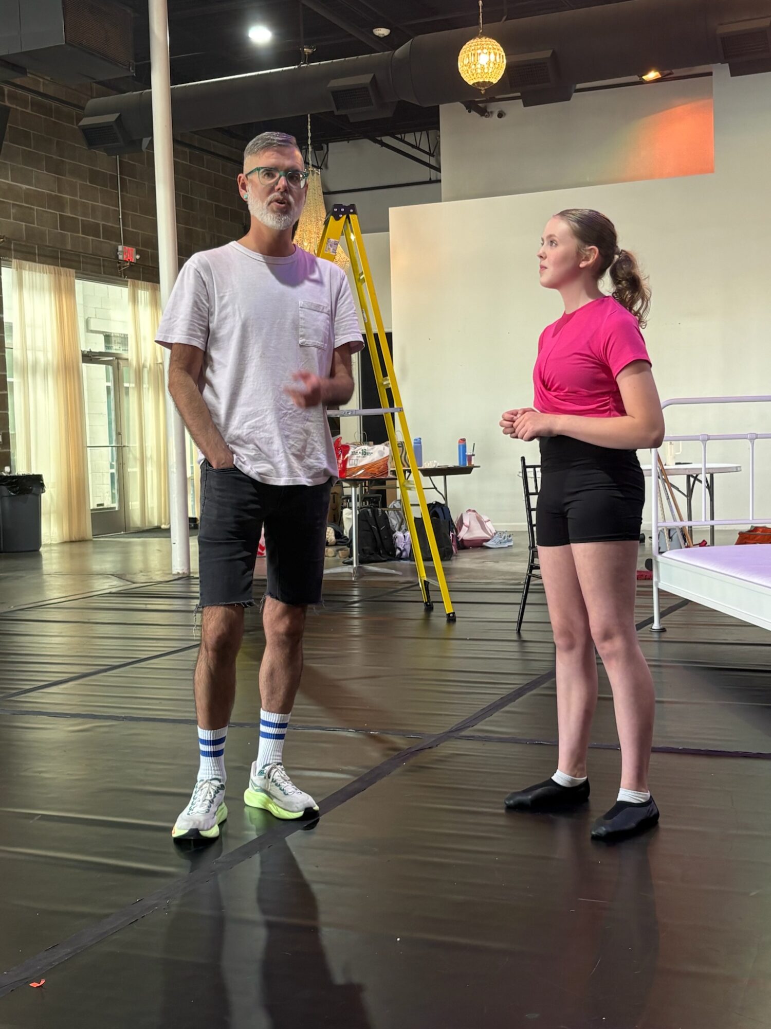 Two people standing and talking indoors, one man and one woman, with a ladder and equipment in the background.