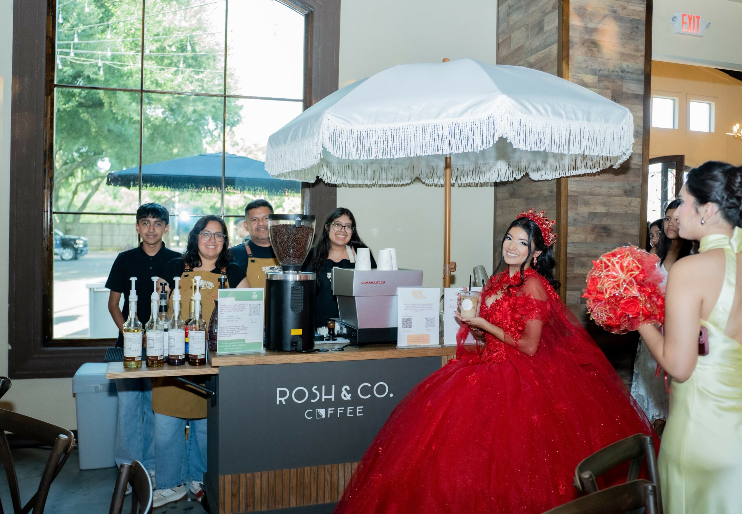 A woman in a red dress holding a bouquet stands inside a cafe with a large window, a counter, and people behind it.