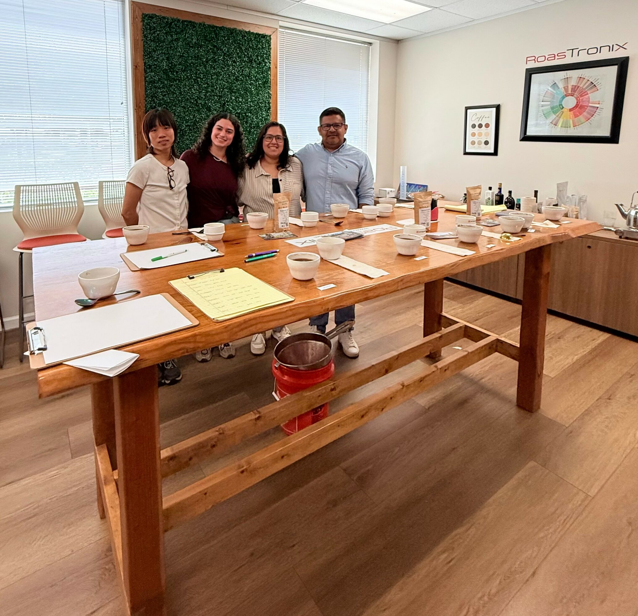 Four people stand behind a long wooden table with art supplies in a bright room with large windows and artwork on the wall.