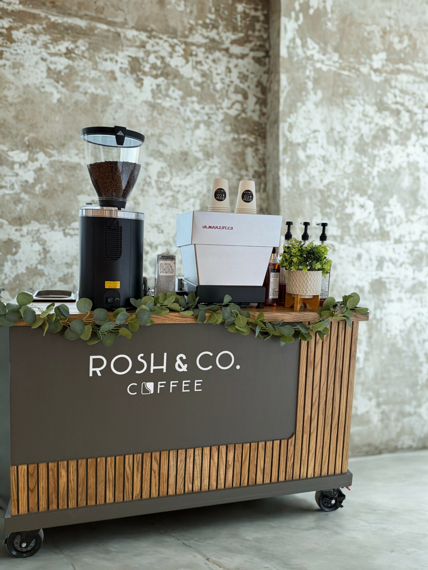 Coffee station with grinder, espresso machine, and plants on a wooden cart against a textured wall.