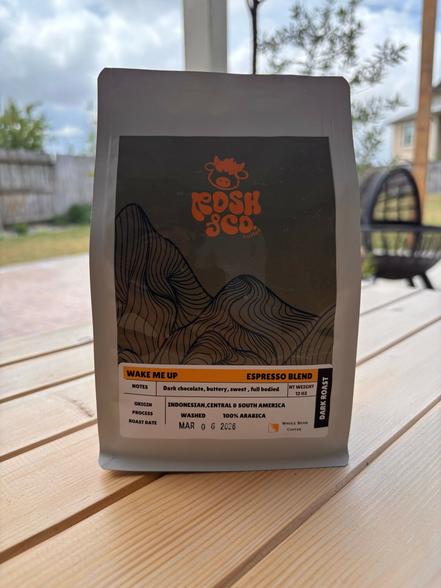 A gray bag with a black label and orange text, placed on a wooden surface near a window with trees outside.