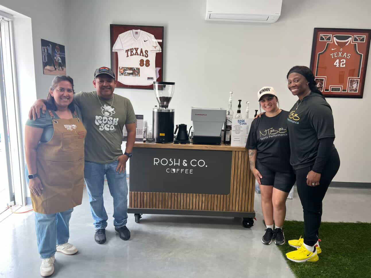 Four people standing behind a coffee counter in a room with sports jerseys on the wall, smiling at the camera.
