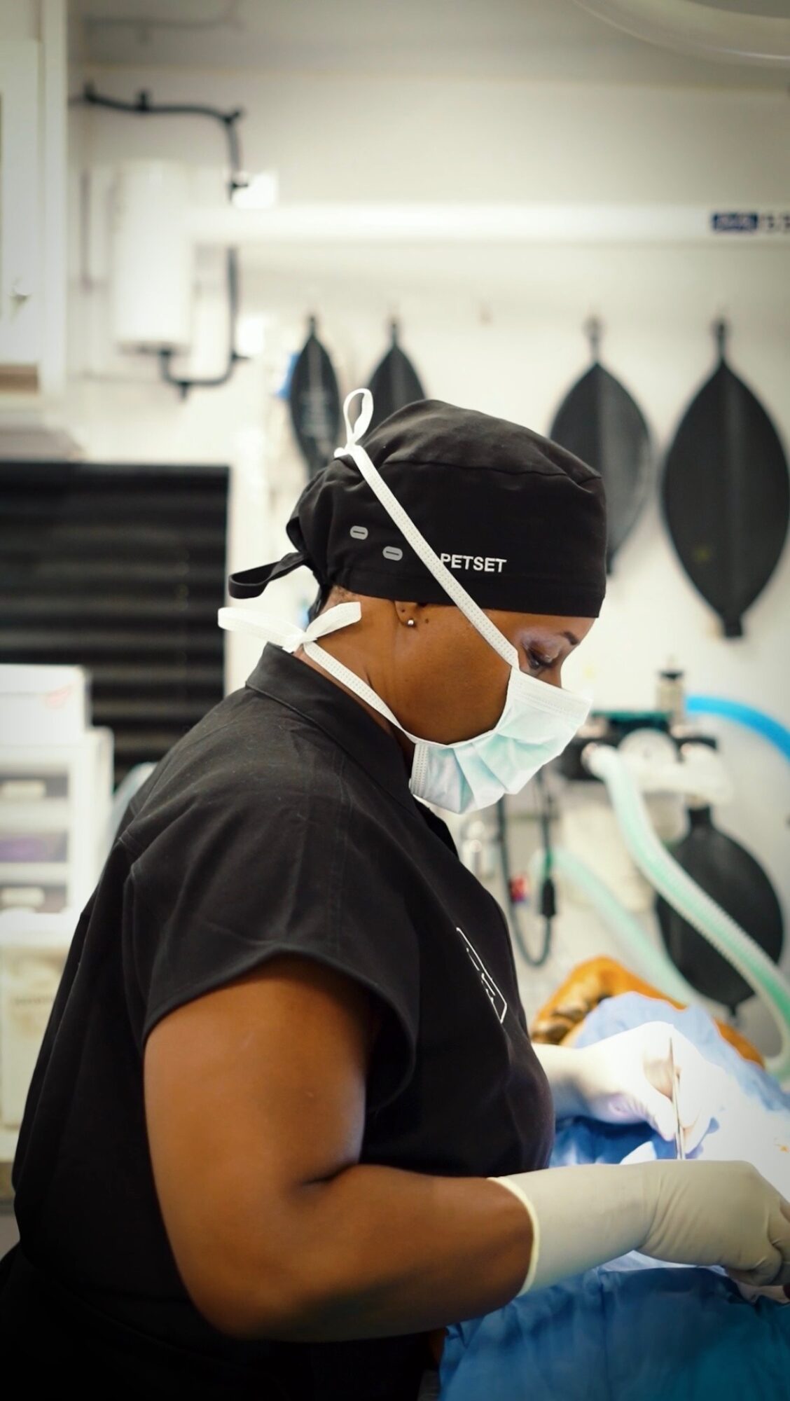 Healthcare worker wearing a mask, gloves, and a black cap, focused on a task in a clinical setting.