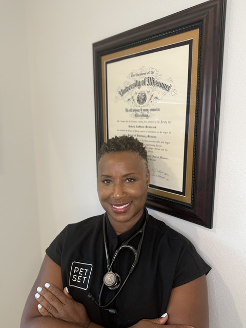 Smiling woman in black scrubs with a stethoscope, standing in front of framed diploma on wall.
