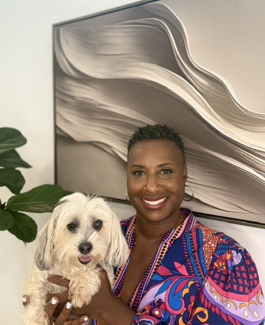 Woman smiling holding a small white dog indoors, with abstract wall art and a plant in the background.