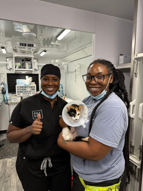 Two women smiling in a laboratory, one holding a small dog with a cone around its neck.