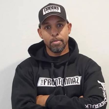 Man wearing a black hoodie and cap with text, standing against a plain white background.