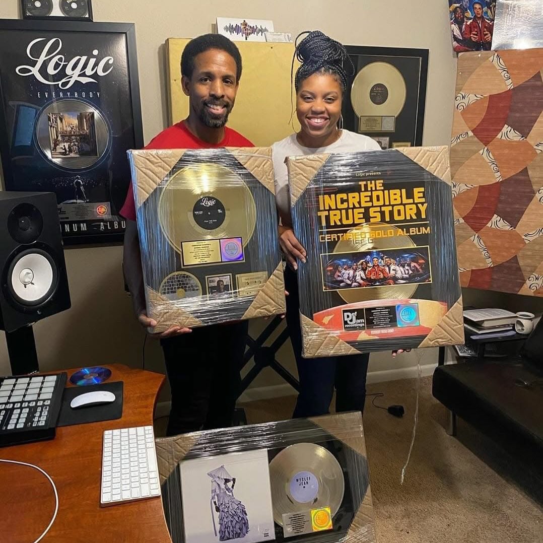 Two people holding framed gold records and a vinyl album in a room with music memorabilia and equipment.