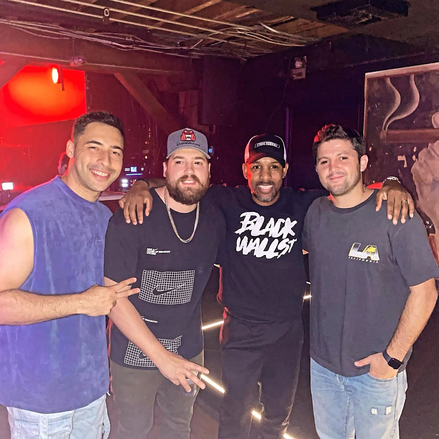 Four men smiling and posing together in a dimly lit indoor setting, with arms around each other, casual clothing, and a red light in the background.