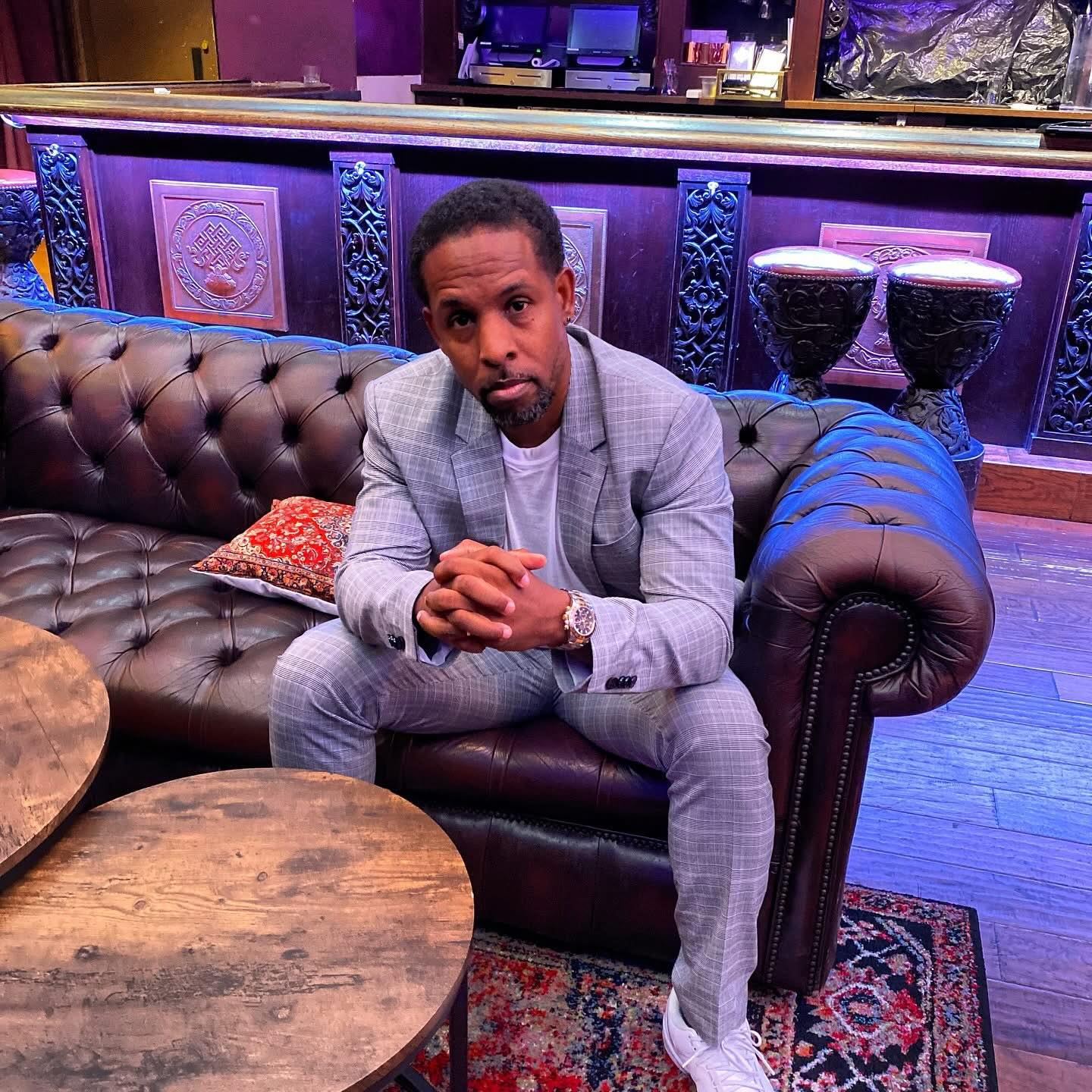 Man in gray suit sitting on a leather sofa with hands clasped, in a room with purple decor and bar stools.