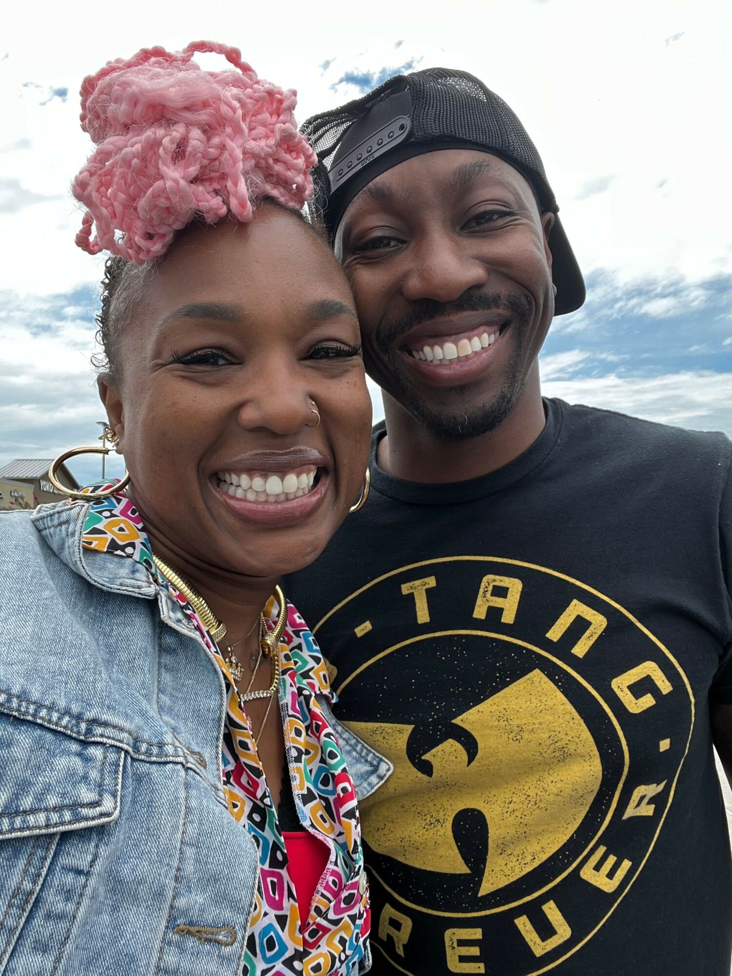 Two smiling people taking a selfie outdoors, one wearing a black cap and the other with pink hair, both smiling broadly.