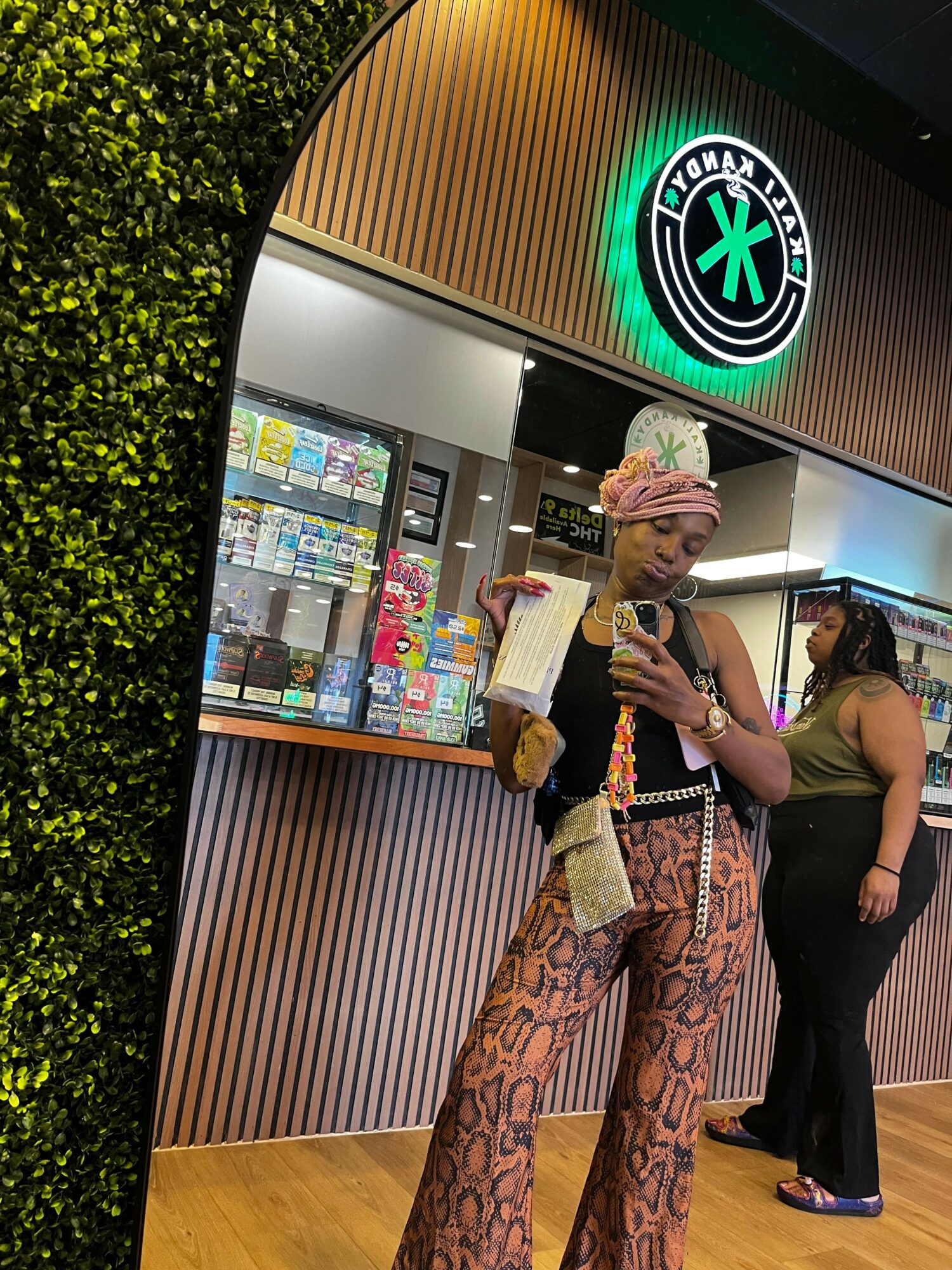 Person in patterned pants and a top holding a drink inside a store with a green logo sign, glass display, and wooden decor.