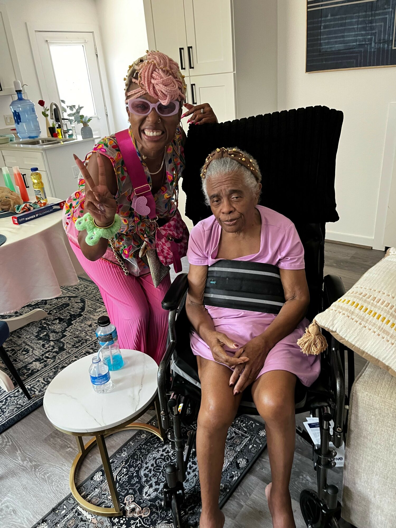 Two women, one in a wheelchair, pose indoors with a mirror and furniture in the background.