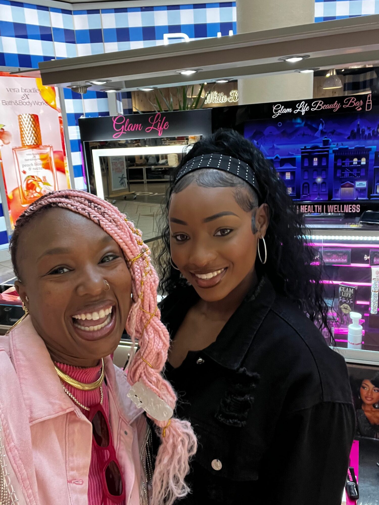 Two women smiling, one wearing a headband and pink jacket, the other with a black jacket and headscarf, in a store.