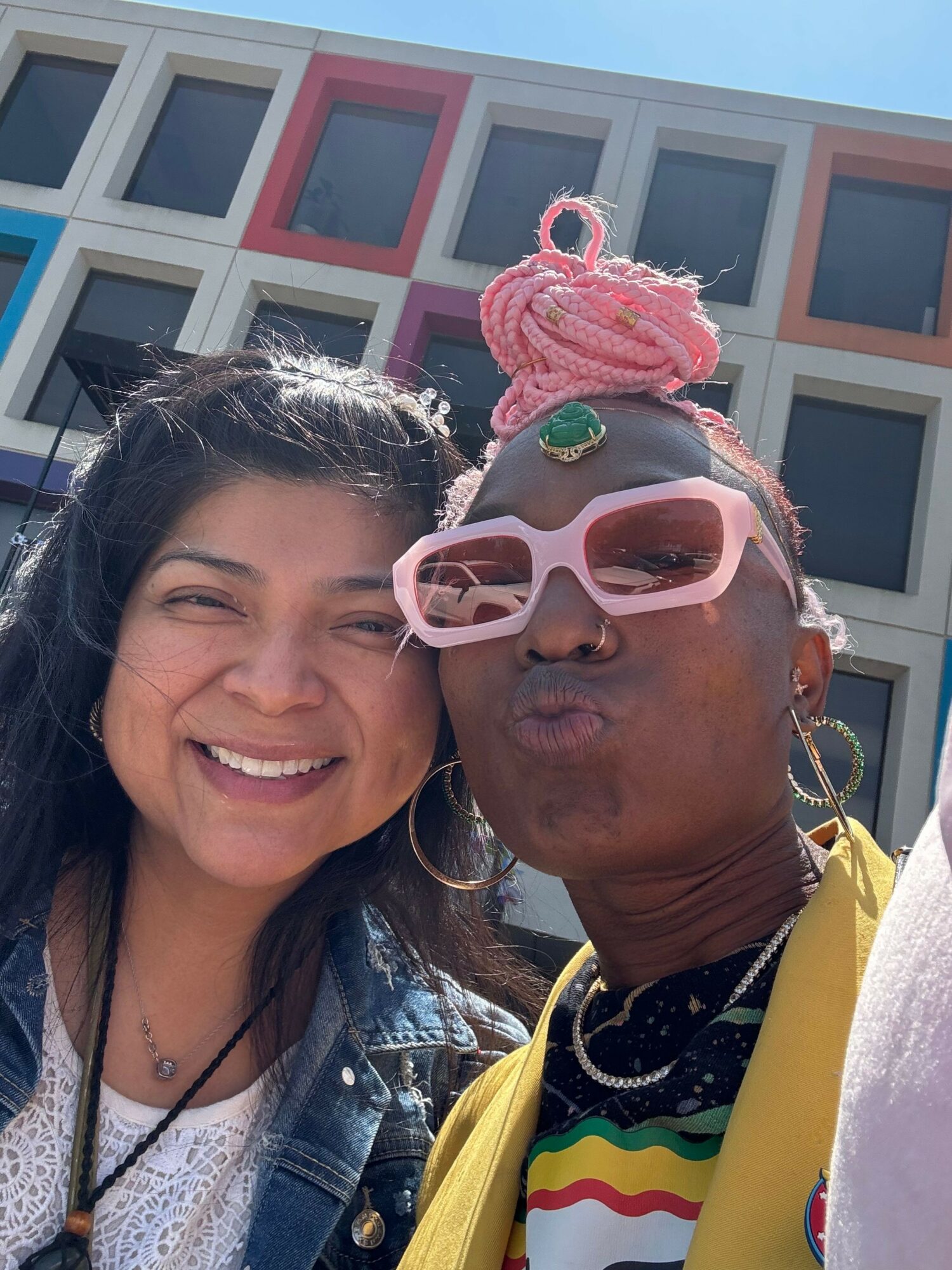 Two people smiling, one wearing pink glasses and a pink wig, the other with long dark hair, in front of a building.