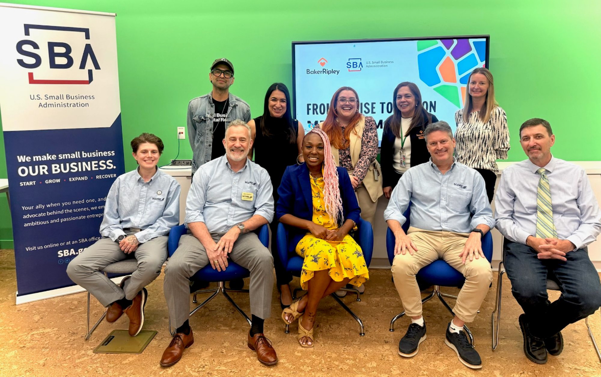 Group of nine people posing in front of a green background and screen, some sitting and some standing, at a professional event.