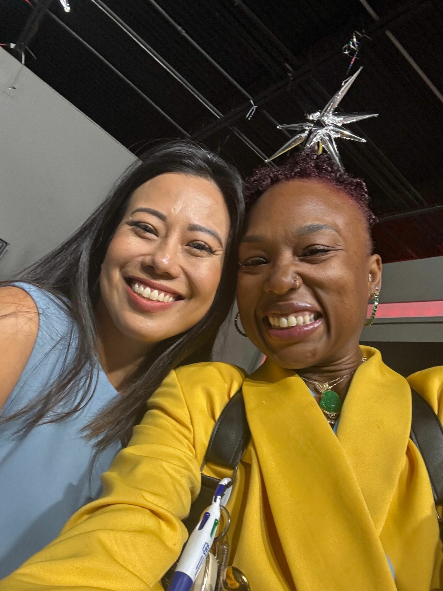 Two women smiling, one wearing a yellow jacket and the other in a light blue top, with a star-shaped decoration in the background.