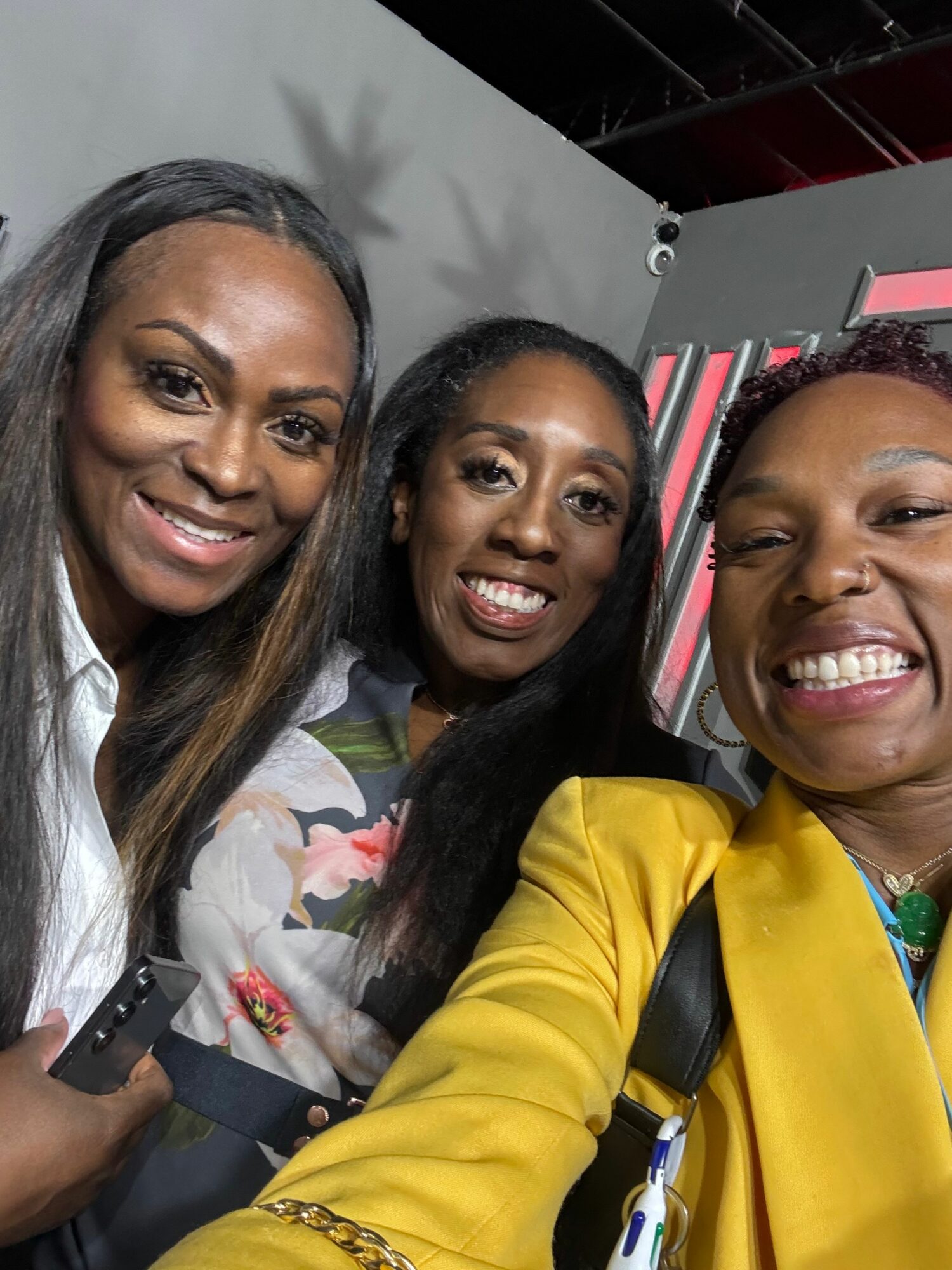 Three women smiling and taking a selfie together indoors, with a gray wall and some pink elements in the background.
