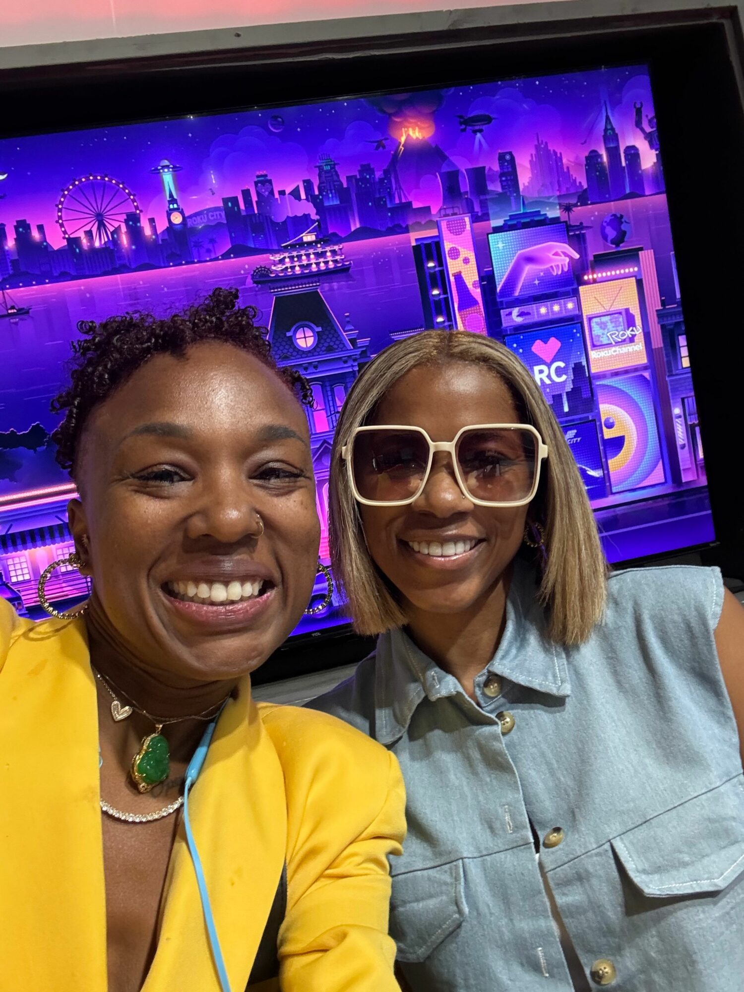Two women smiling in front of a colorful, purple-lit stage or event backdrop, one wearing large glasses and the other in a yellow jacket.