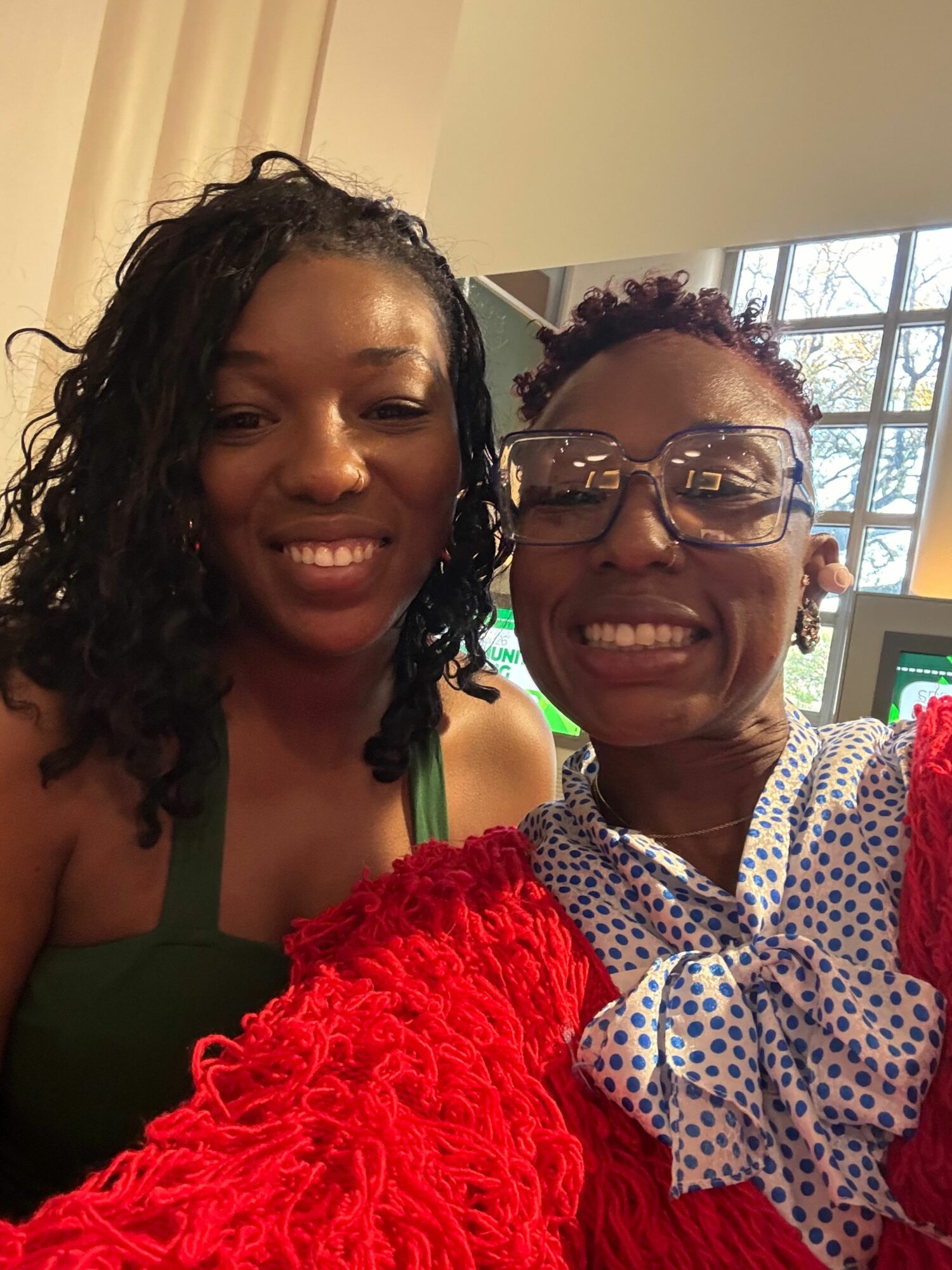 Two women smiling, one wearing glasses and a polka-dot shirt, the other with curly hair and a green top, indoors.