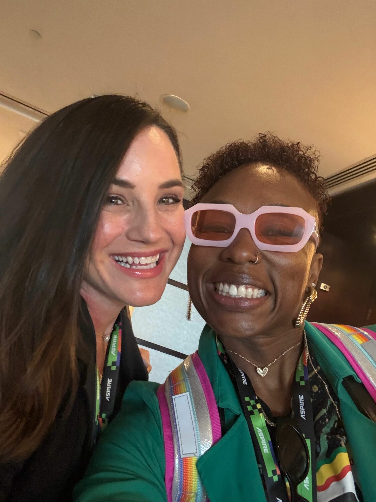 Two women smiling, one wearing pink sunglasses, the other with long dark hair, indoors with a beige wall background.