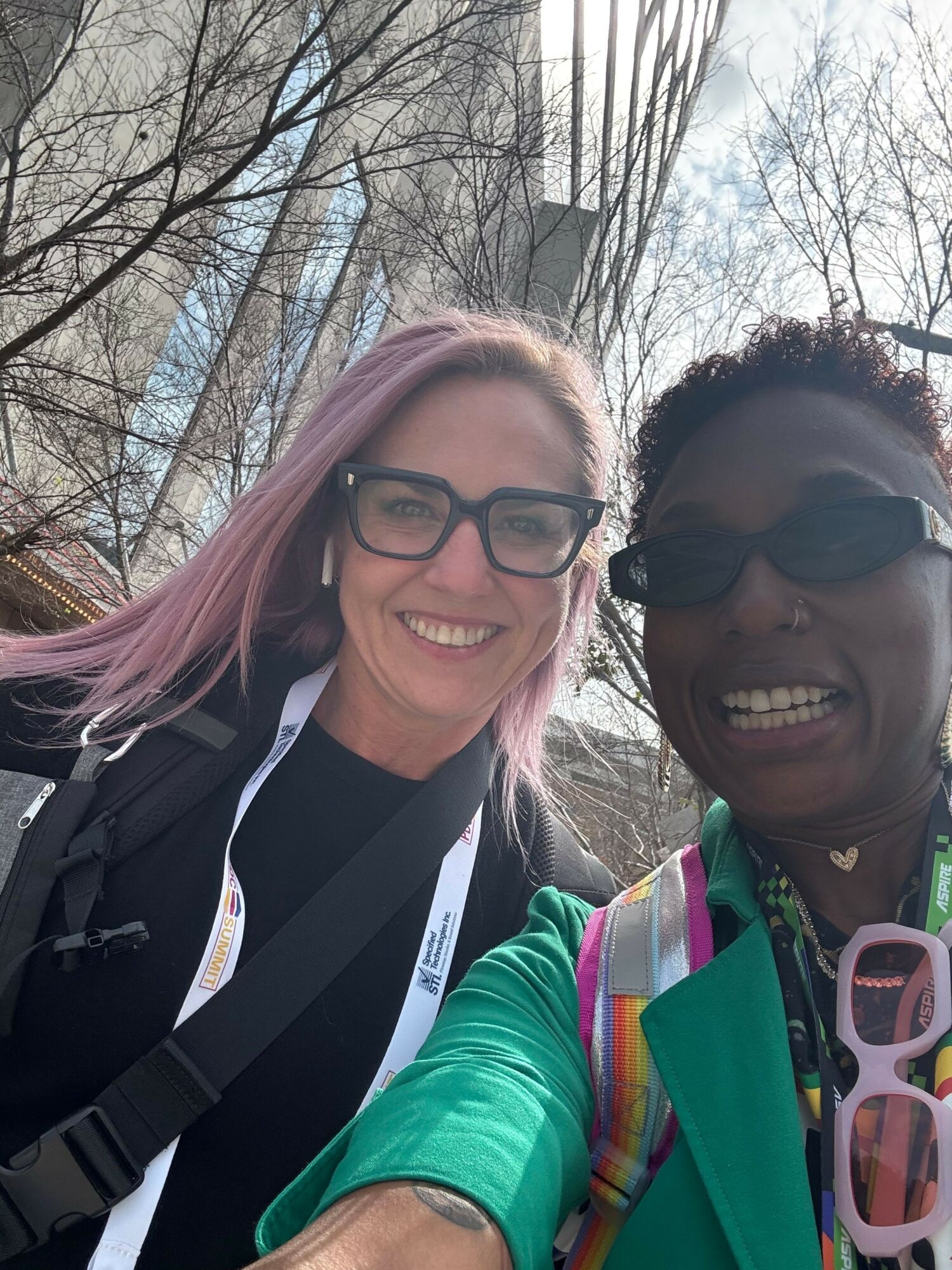 Two smiling women outdoors, one with pink hair and glasses, the other with dark hair and sunglasses, trees in background.