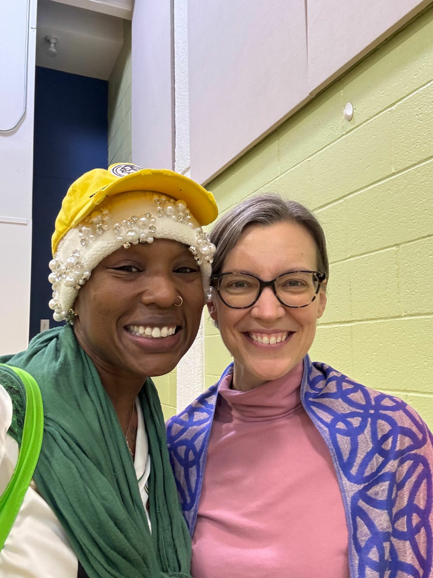 Two women smiling, one wearing glasses and a pink shirt, the other with a yellow headscarf and white headwrap, indoors.