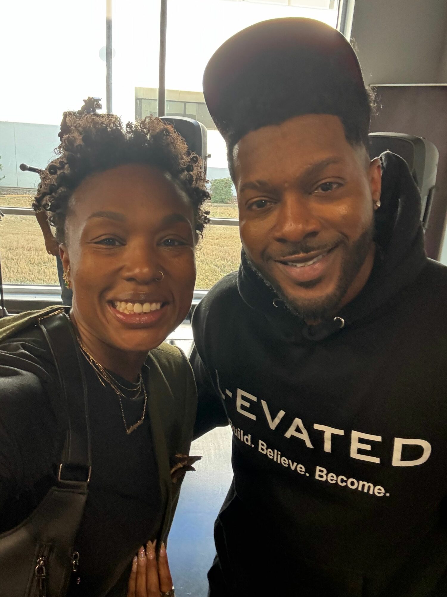 Two people smiling, one wearing a black hoodie and the other a black jacket, standing outdoors near a window.