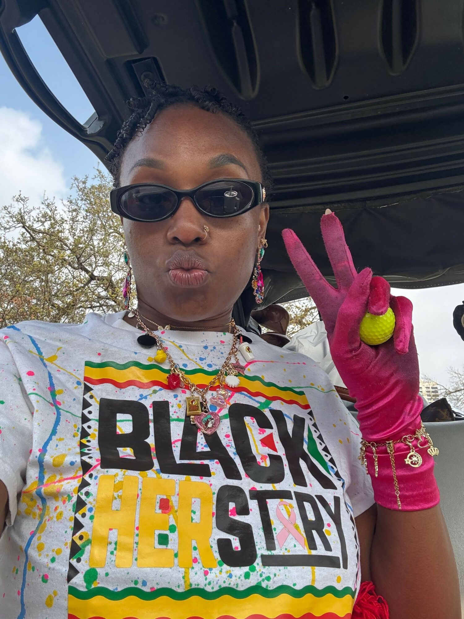 Person wearing sunglasses and a colorful shirt making a peace sign with pink gloves, holding a tennis ball.
