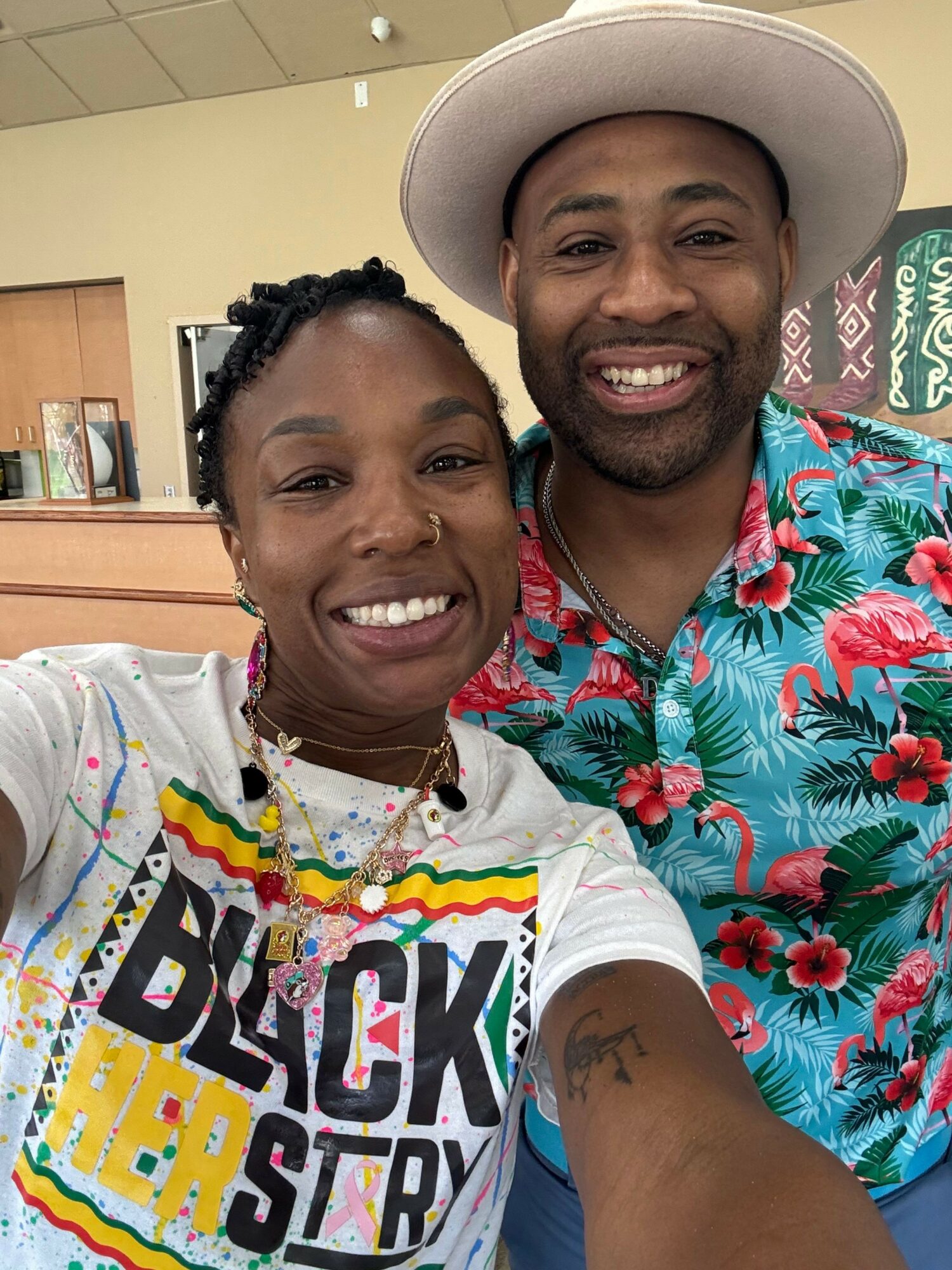 Two smiling people taking a selfie, one wearing a floral shirt and hat, the other in a colorful graphic T-shirt.