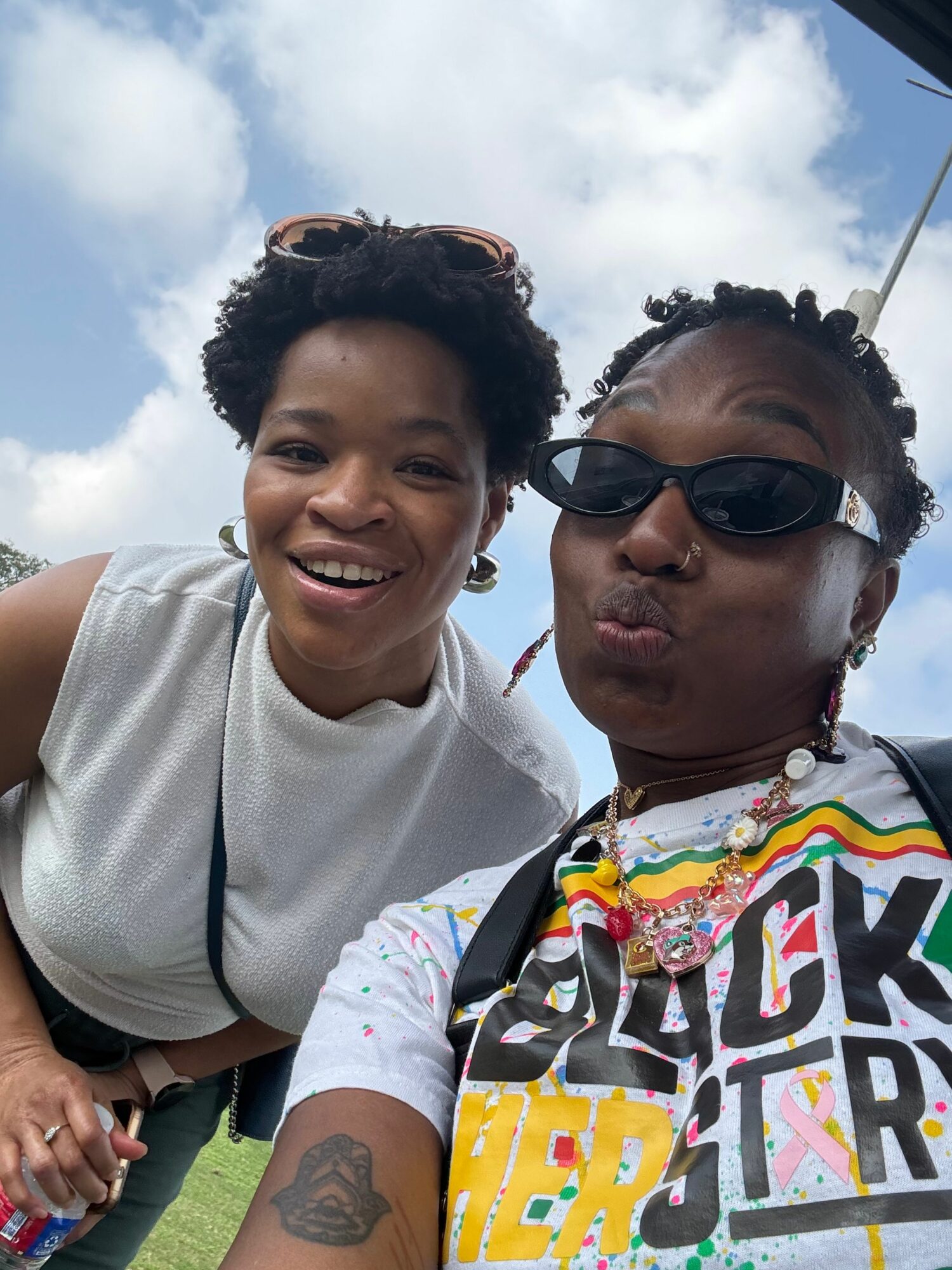 Two women smiling outdoors, one wearing sunglasses and a colorful shirt, the other in a white top, with a cloudy sky background.