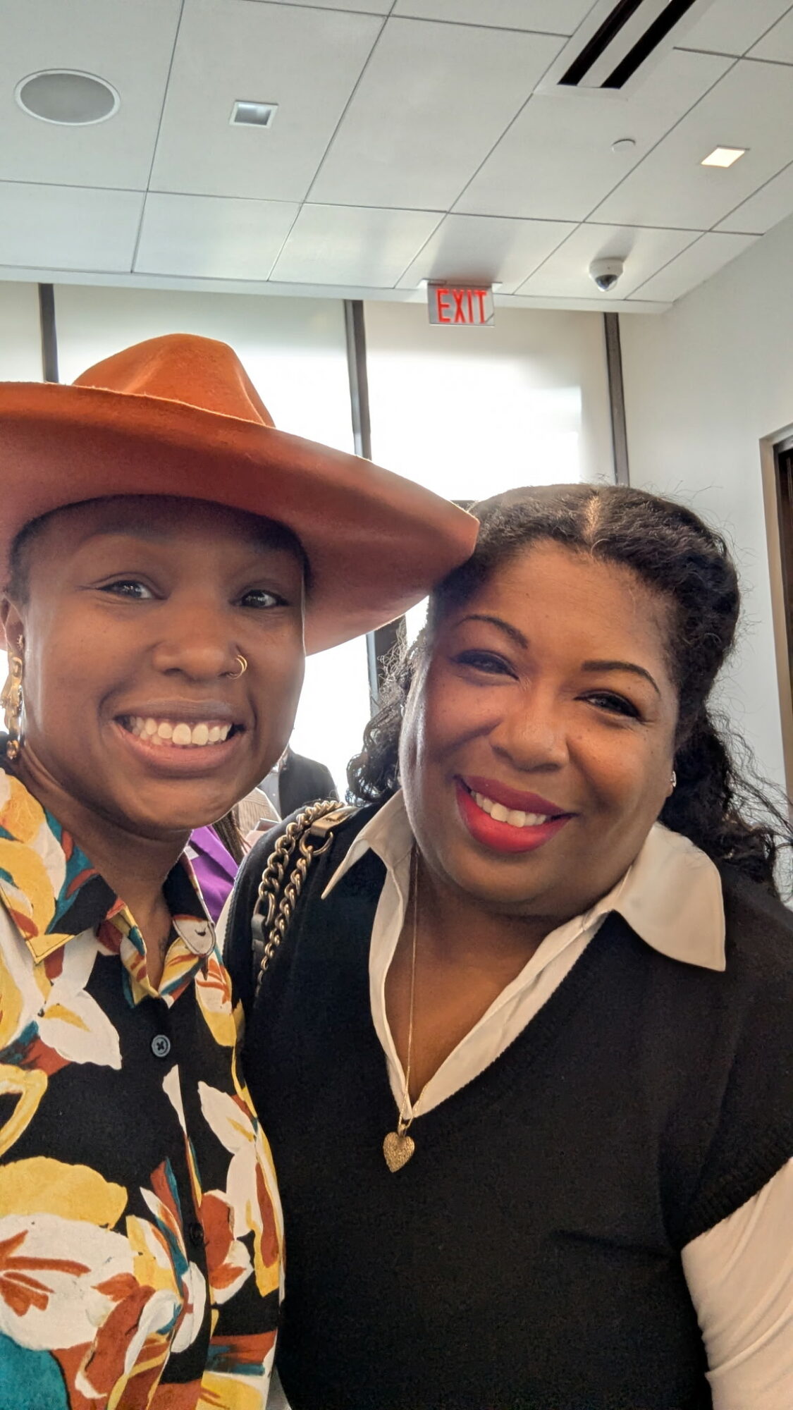 Two women smiling indoors, one wearing a large hat and colorful patterned shirt, the other in a black and white outfit.