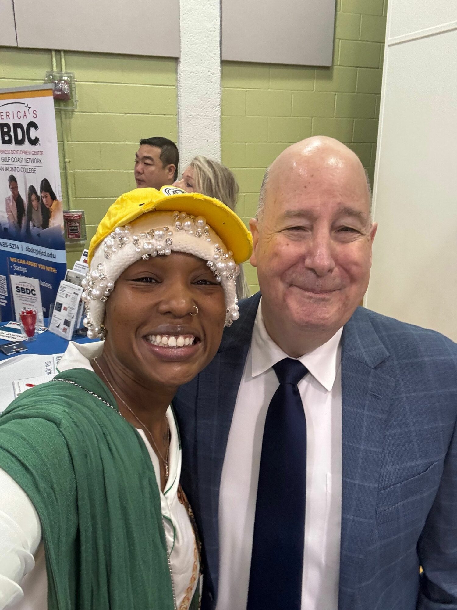 Two people smiling, one wearing a yellow hat and green shirt, the other in a suit, in an indoor setting.