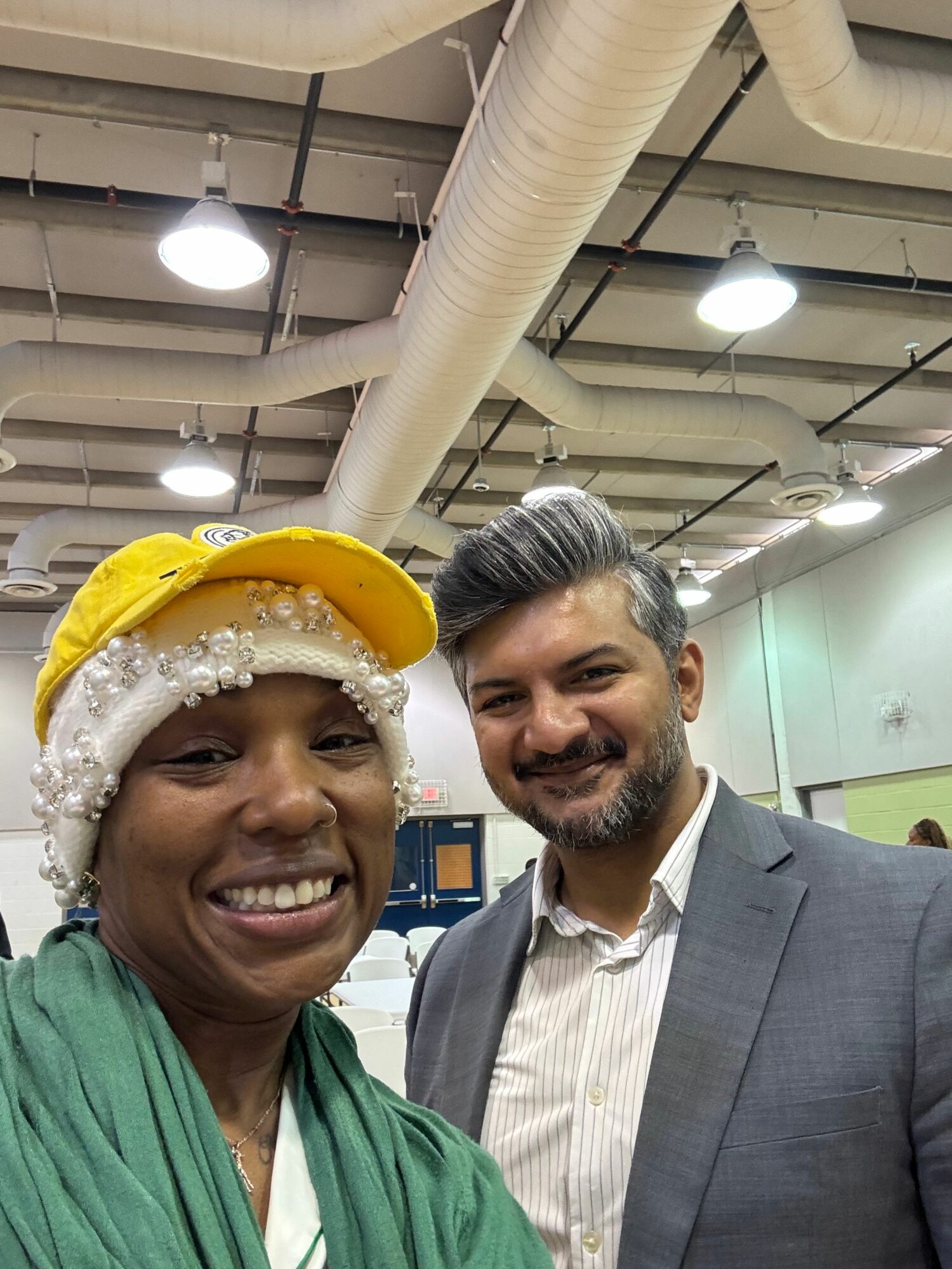 Two smiling women, one wearing a yellow hat and green scarf, the other in a suit, in an indoor setting.