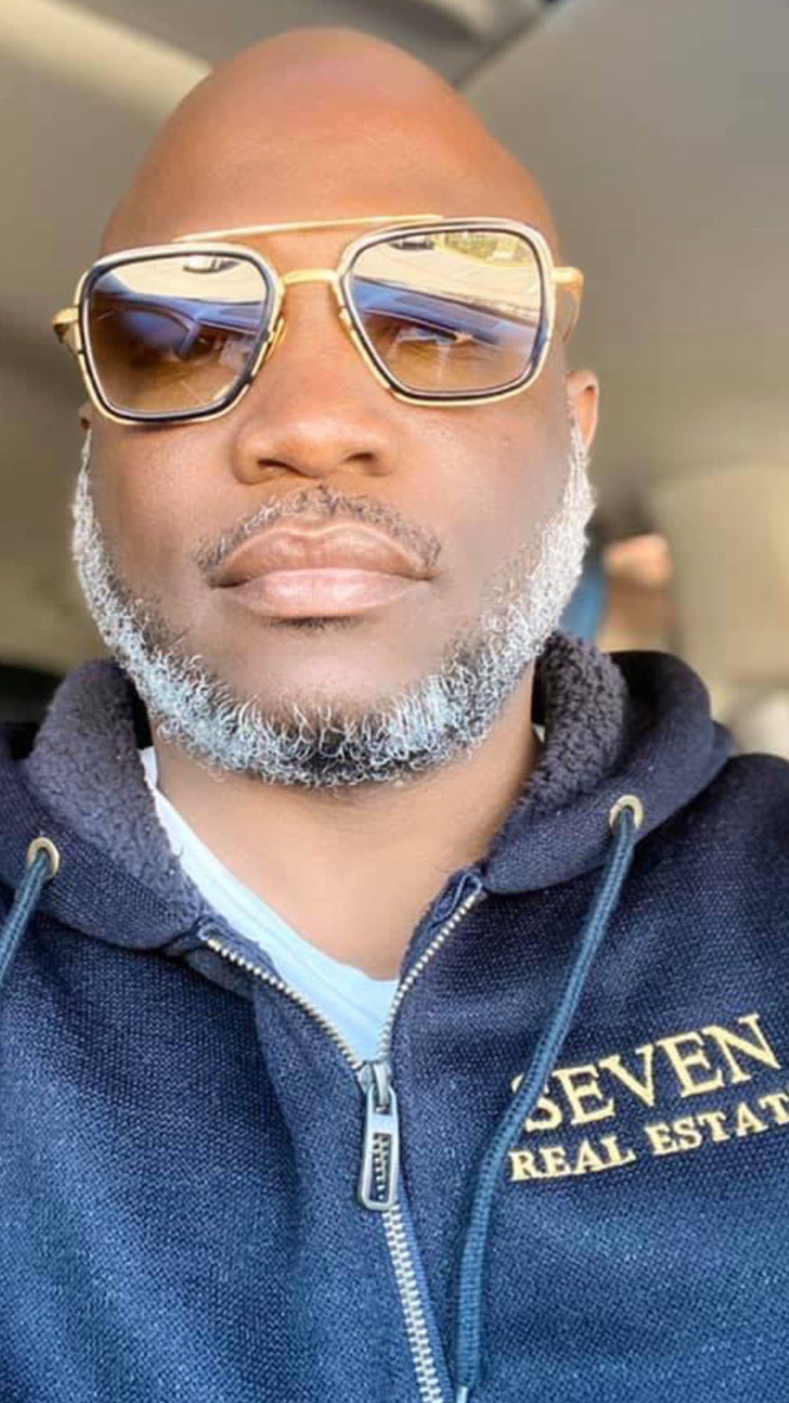 Bald man with sunglasses and gray beard wearing a navy hoodie with gold lettering, inside a vehicle.