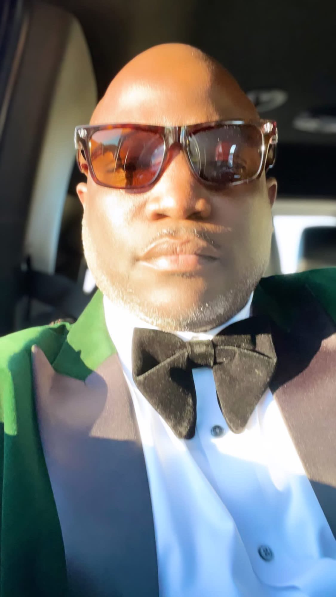 Person wearing sunglasses and a tuxedo with a bow tie, sitting in a vehicle, looking at the camera.