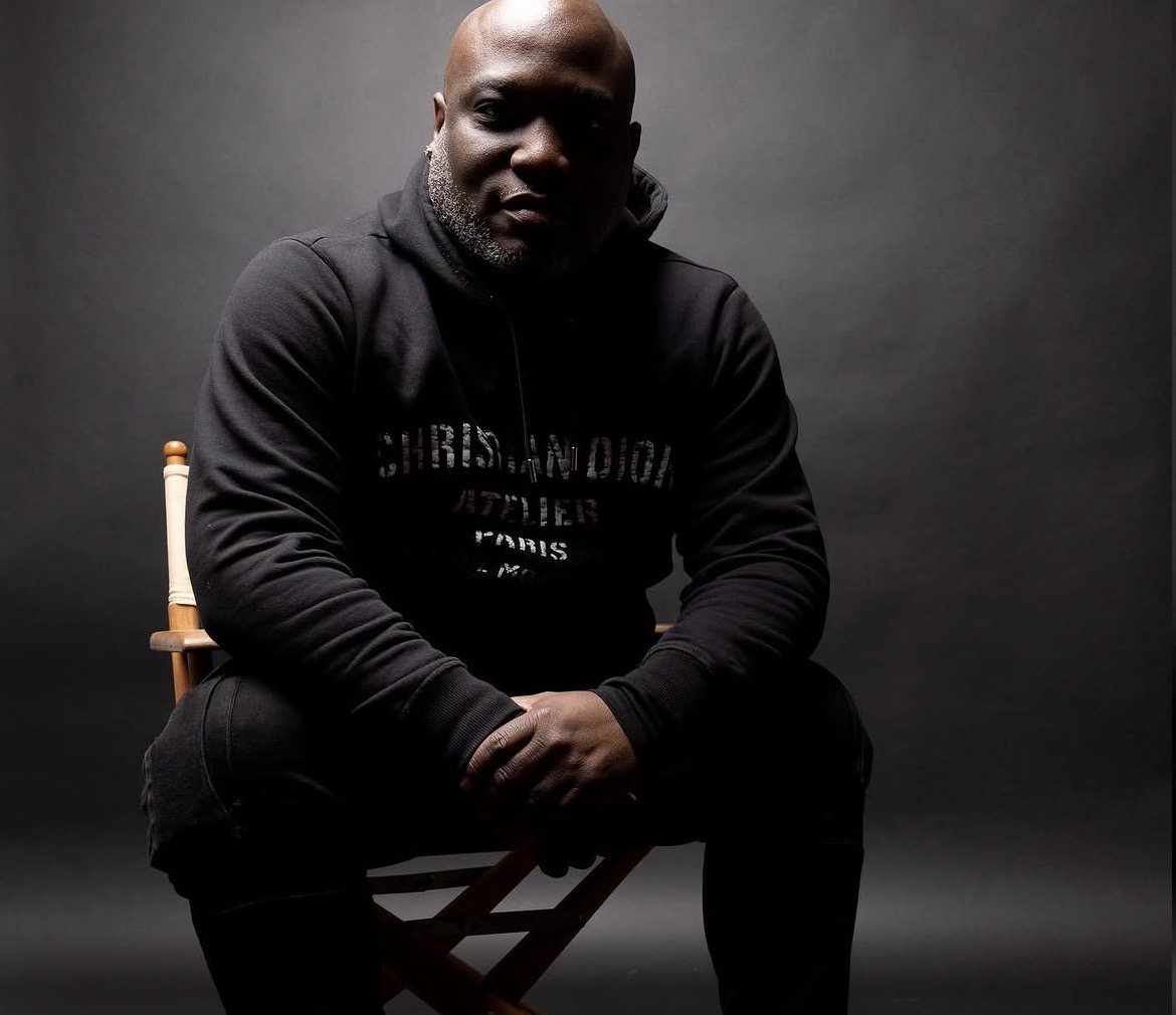 Person sitting on a chair against a dark background, wearing a black hoodie, with hands clasped, looking at the camera.