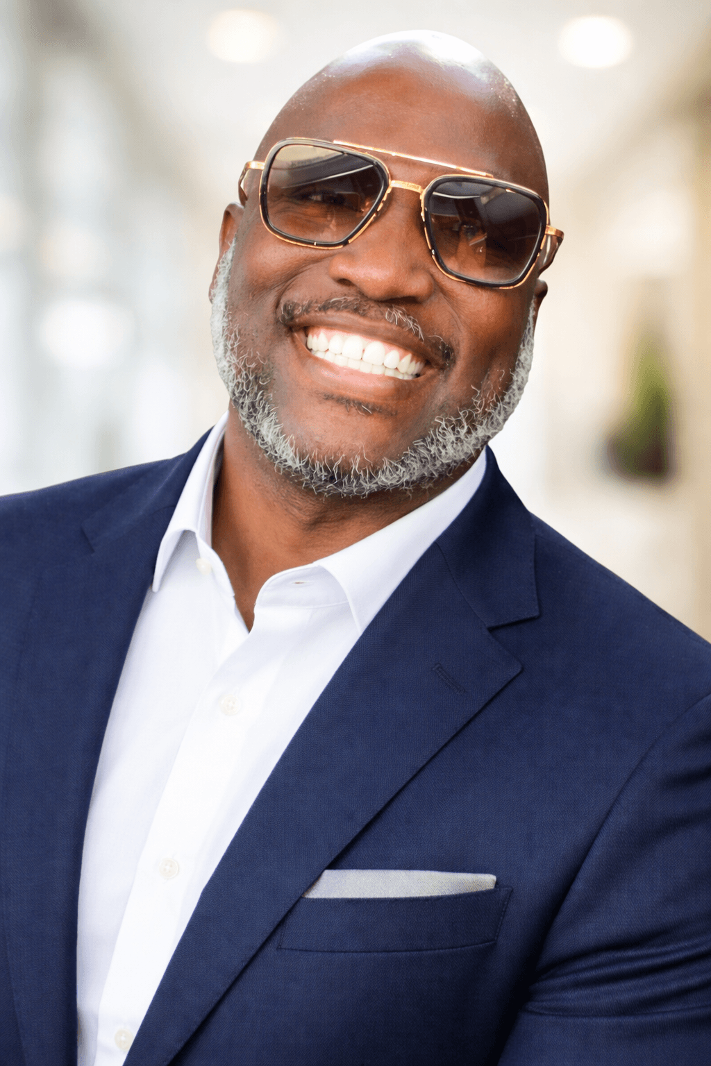 Smiling man wearing sunglasses, a white shirt, and a navy blazer outdoors with blurred background.
