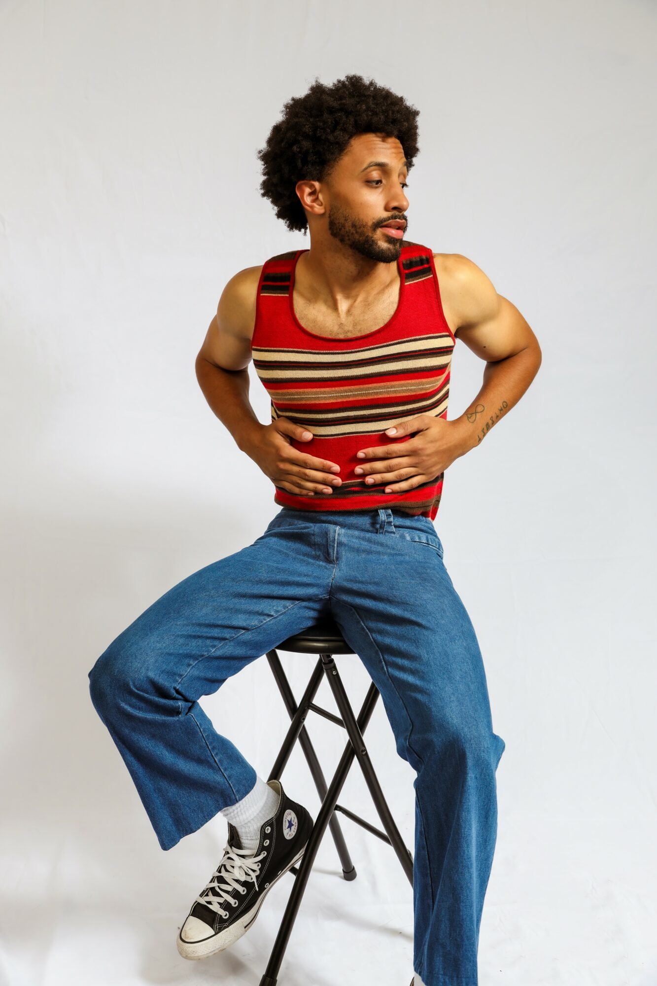 Person with curly hair and beard sitting on a stool, wearing a striped tank top and jeans, looking to the side.