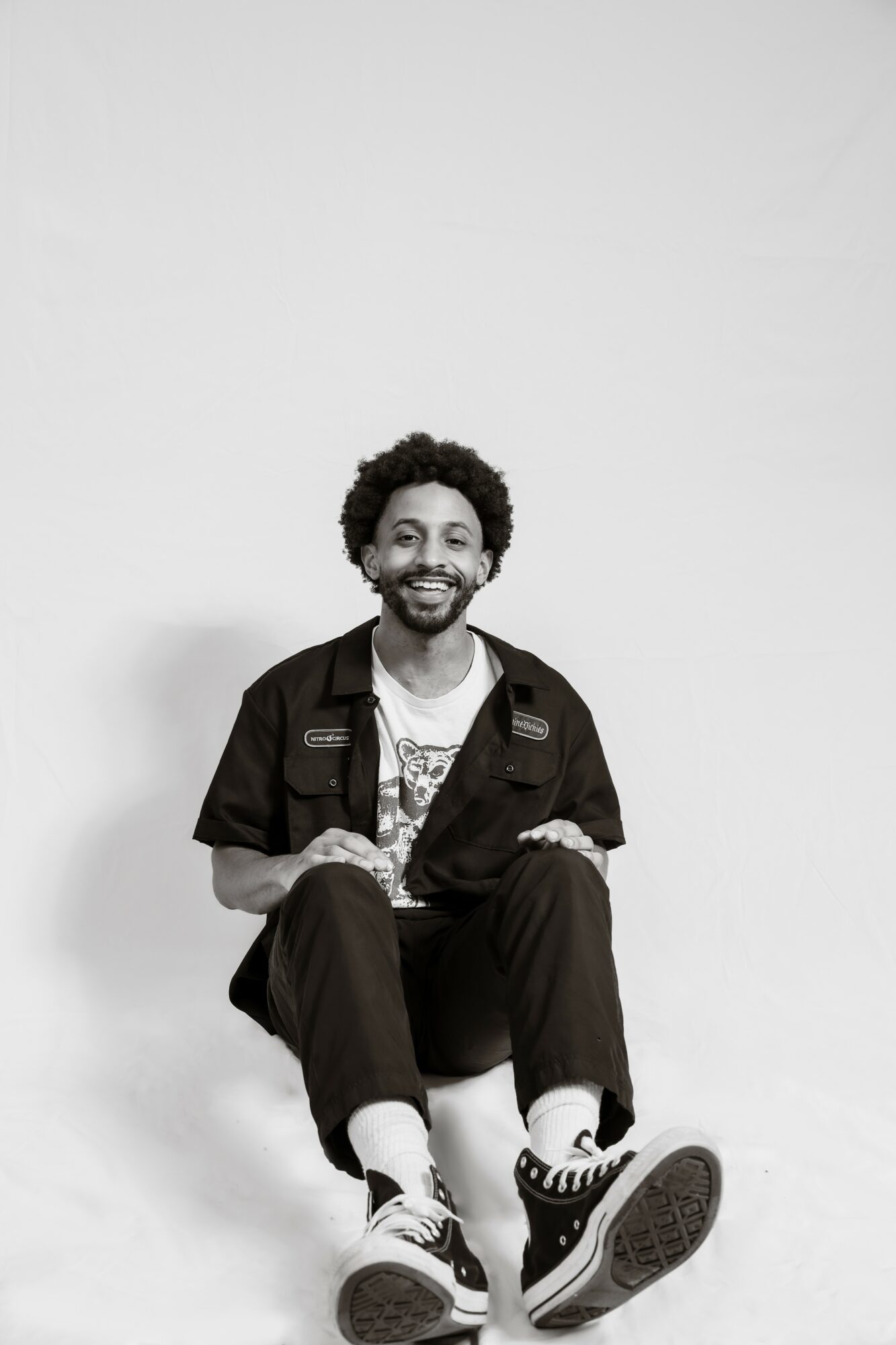 Person with curly hair and beard sitting on a white surface, smiling, wearing a dark shirt and pants, sneakers, against a plain background.