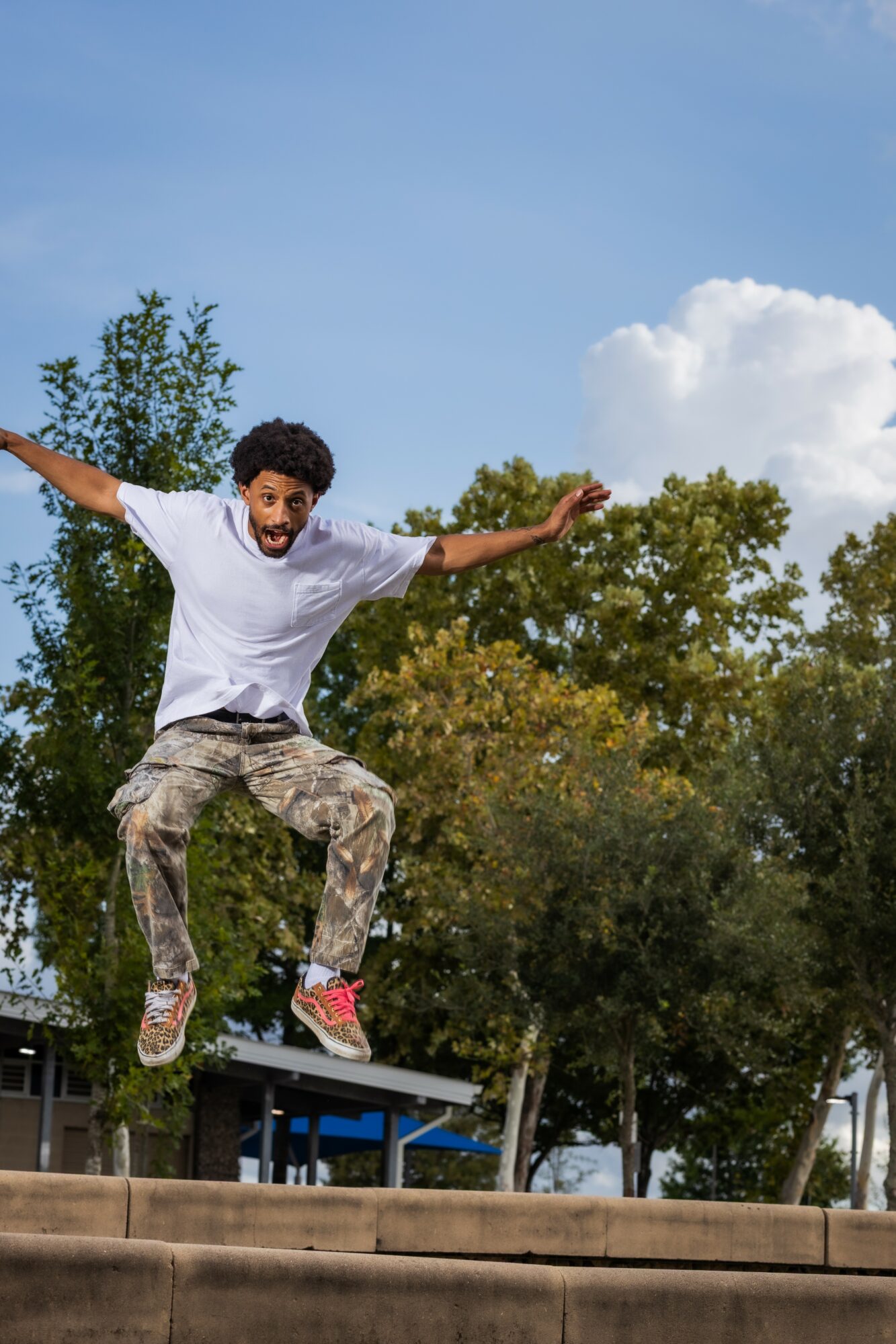 Person jumping with arms outstretched outdoors, trees and blue sky in background.