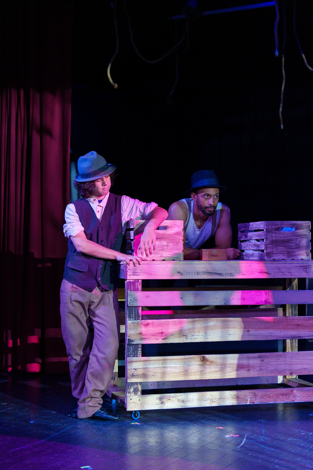 Two men in hats and vests work with a wooden structure on stage, with dark background and stage lighting.