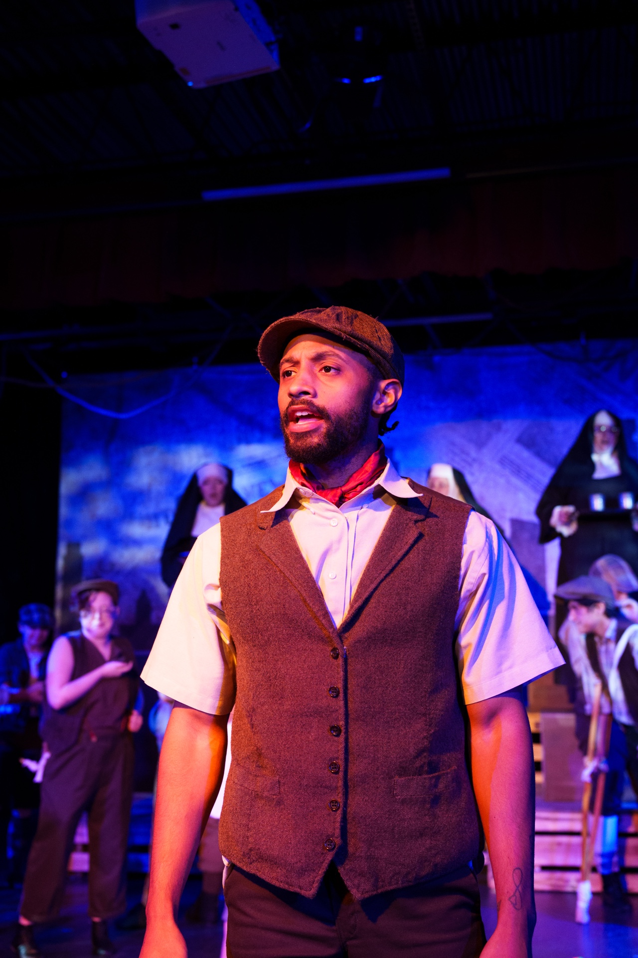Man with beard and cap on stage, wearing vest and shirt, with people in background.