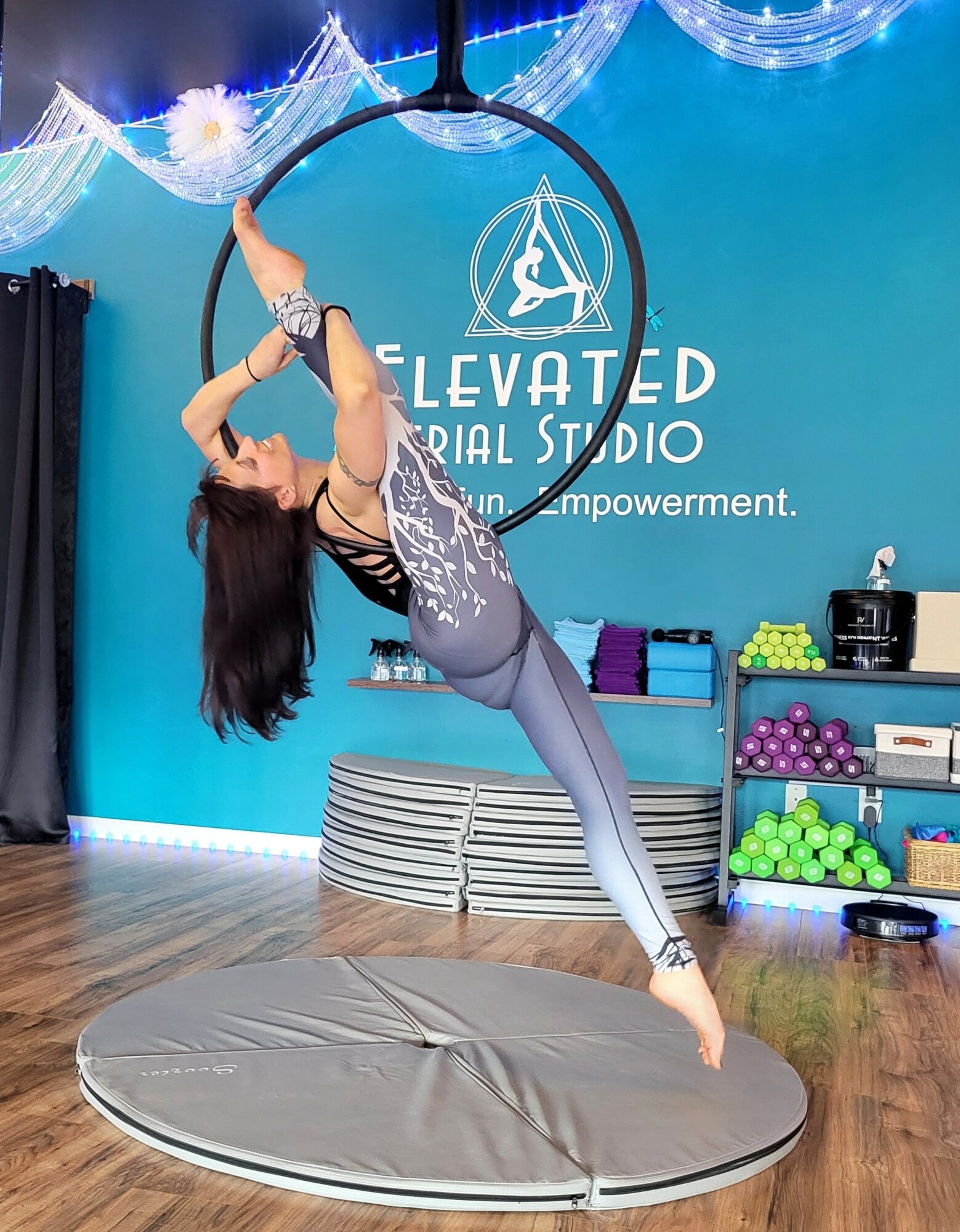A woman performs aerial acrobatics on a hoop in a studio with blue walls and equipment, hanging upside down with one leg extended.