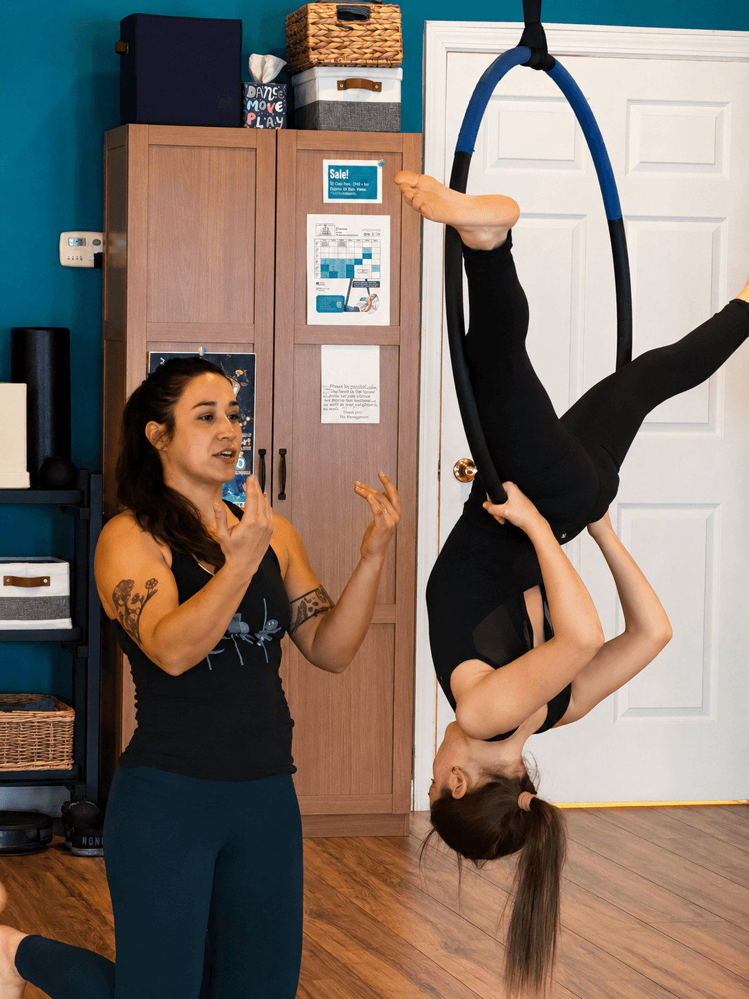 A woman performs aerial acrobatics on a hoop, while another woman watches and gestures indoors.
