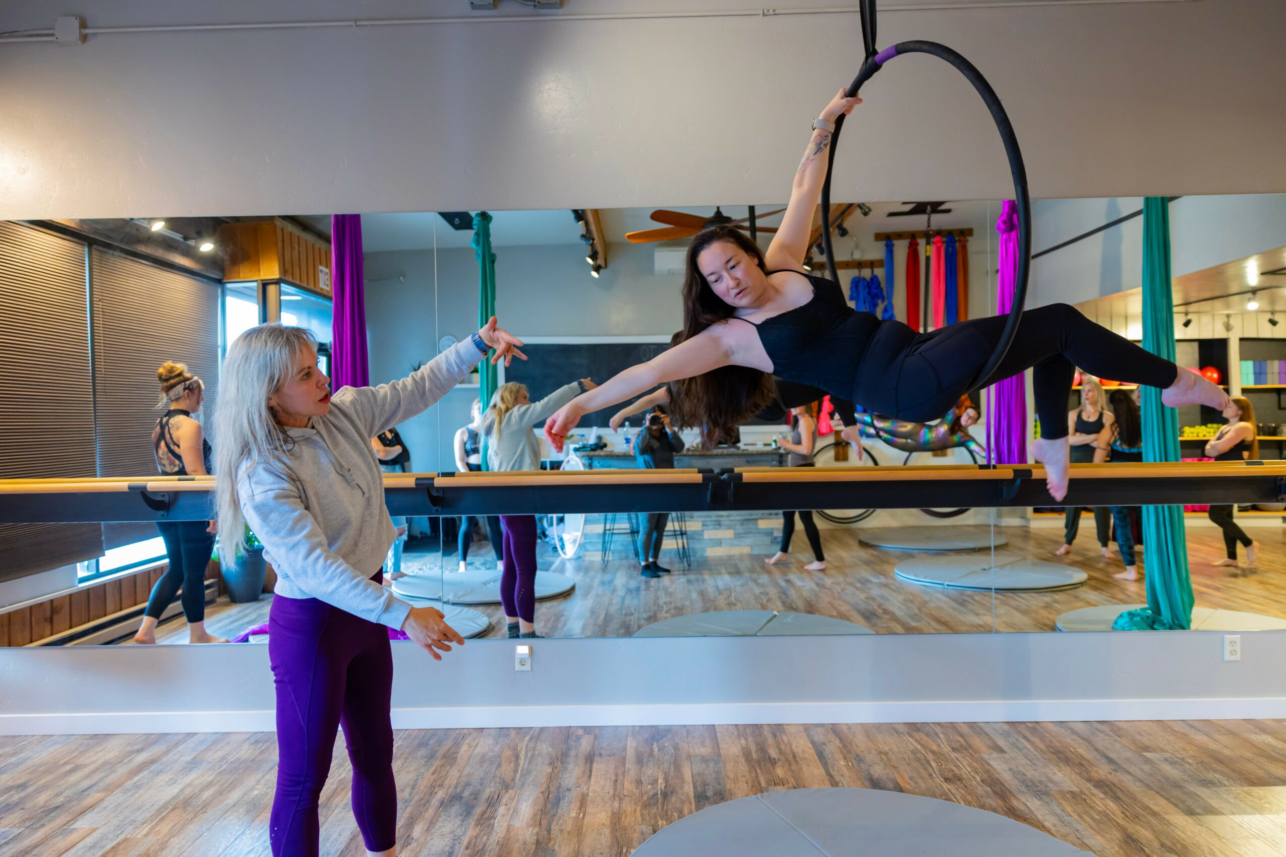 Woman performing aerial hoop exercise in a fitness studio with mirrors and other people in the background.
