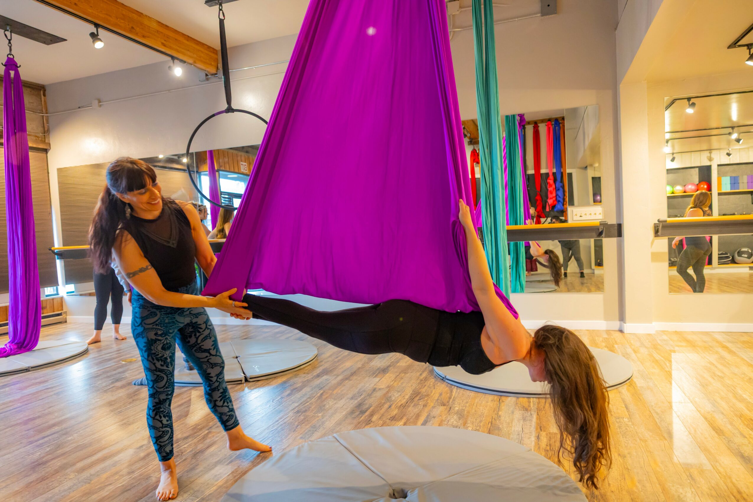 Two people in an indoor space with colorful hanging fabric, one holding a purple fabric swing and the other lying on it.