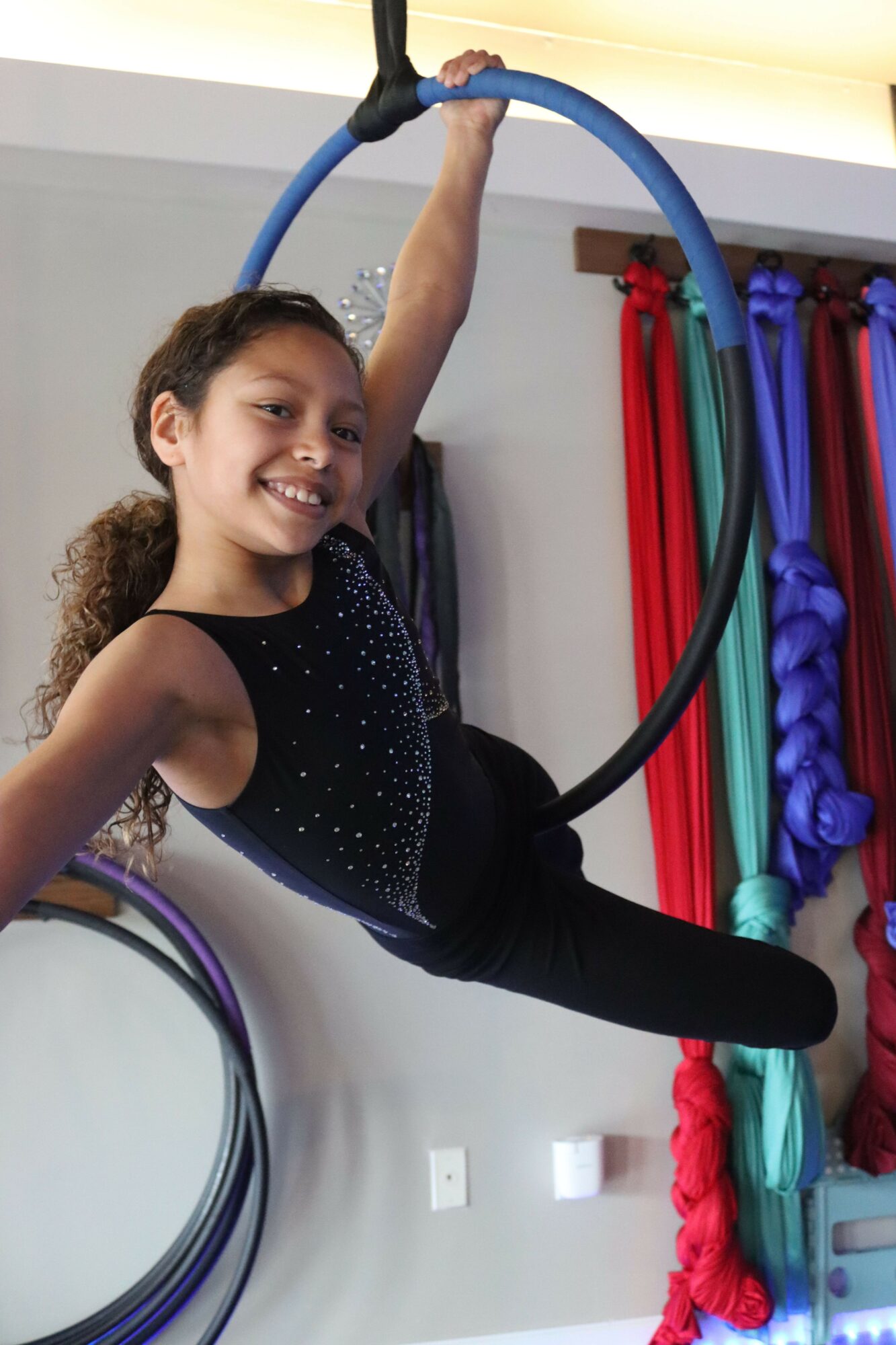 Young girl performing a hoop dance, smiling, in a room with colorful fabric rolls on shelves.