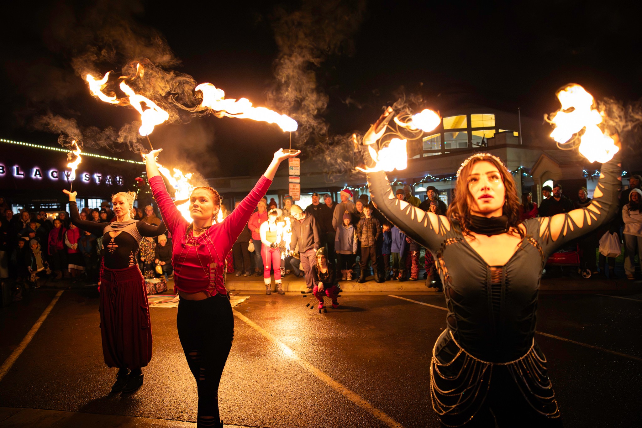 Two fire performers hold flaming torches with a crowd watching at night, illuminated by firelight, in an outdoor event.